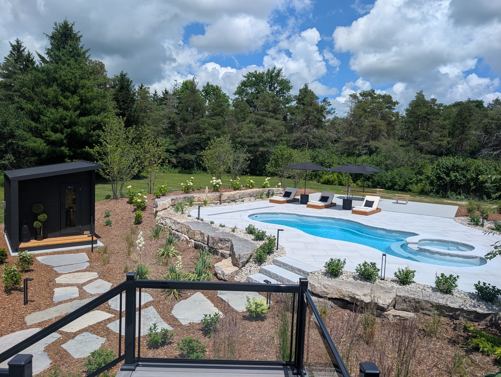 Curving fibreglass pool below a stone retaining wall and garden, with loungers and a cube sauna