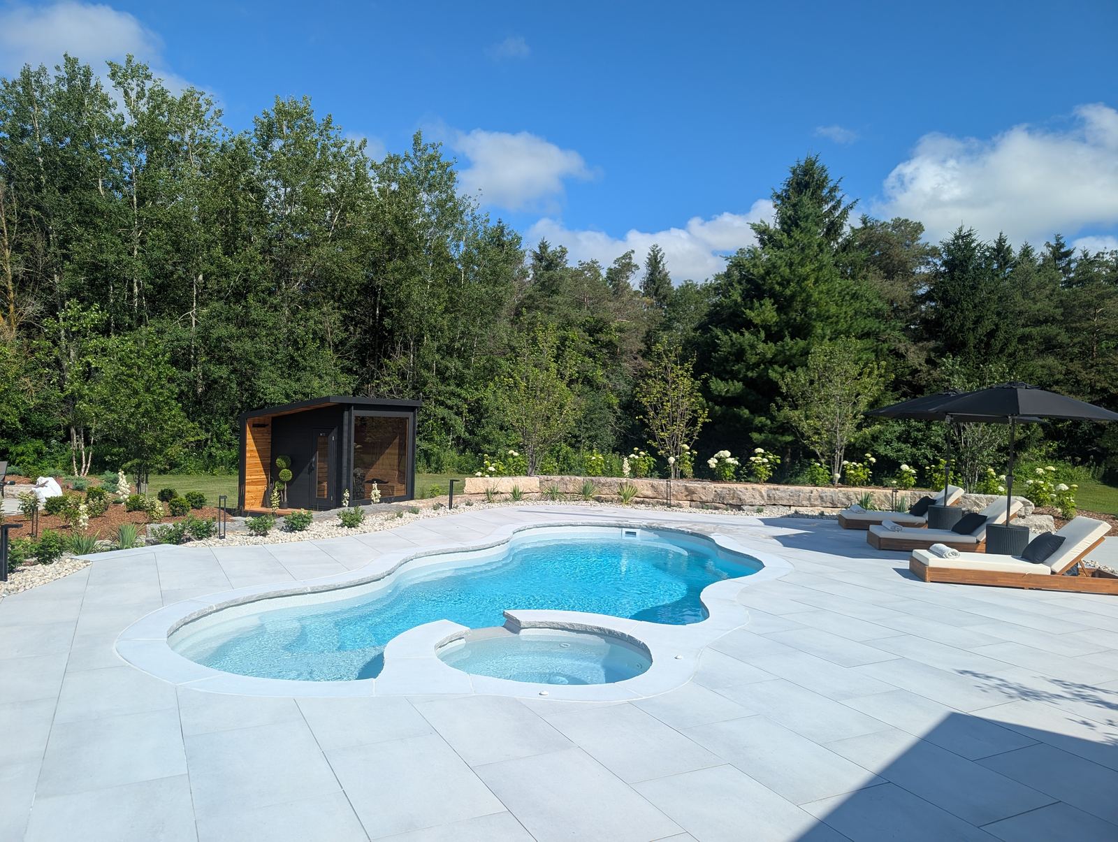 Freeform fibreglass pool with attached spa, stone patio and a black cube sauna in a treed backyard